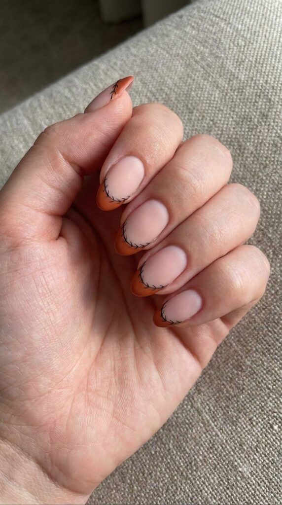 Short almond nude nails with burnt orange French tips and black stitches in matte